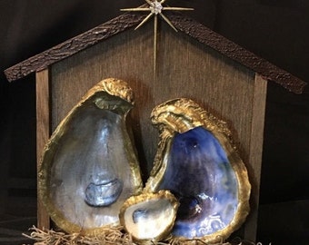 May include: A handcrafted nativity scene featuring oyster shells with gold trim, arranged within a wooden stable. The shells are painted in shades of silver and blue, with a small shell representing the baby. A star tops the stable.