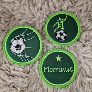 Könnte beinhalten: Drei Fußball-Patches. Einer zeigt einen Ball im Netz, ein anderer einen Fußballspieler und der dritte den Namen "Marinus" mit einem Stern. Alle haben einen grünen, gezackten Rand.