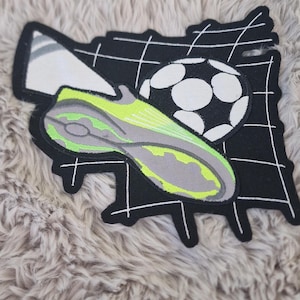Könnte beinhalten: Ein gestickter Aufnäher mit einem lindgrünen und grauen Fußballschuh vor einem schwarz-weißen Fußball und einer weißen Tornetzgrafik. Das Design ist mit schwarzem Garn umrandet und hat einen weißen Rand auf schwarzem Hintergrund.