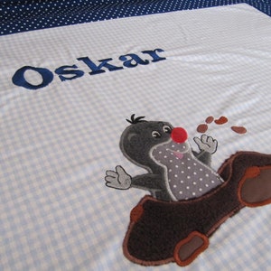 Könnte beinhalten: Eine blaue und weiße karierte Babydecke mit dem Namen "Oskar" in blauer Stickerei. Die Decke zeigt einen gestickten Comic-Maulwurf mit roter Nase, braunem Fell und weißen Punkten. Der Maulwurf schaut aus einem braunen Baumstamm.