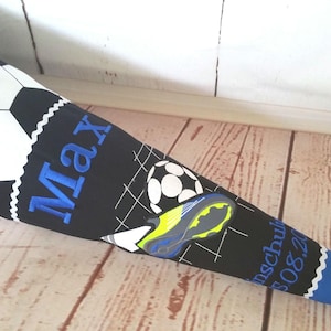 Könnte beinhalten: Eine blaue und schwarze Schultüte mit Fußball-Design. Der Kegel zeigt den Namen "Max" in blauer Stickerei, zusammen mit einem Fußball und einer Fußballschuh-Grafik. Der Kegel ist aus Stoff und hat eine spitz zulaufende Form.