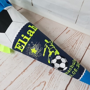 Könnte beinhalten: Eine blau-weiße Schultüte mit Fußball-Design. Auf der Tüte steht der Name "Elijah" in Neongrün, zusammen mit einer Fußballspieler-Grafik und einem Fußball. Die Tüte ist mit Sternen und einem Datum verziert.