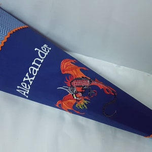 Könnte beinhalten: Ein blauer und orangefarbener Schultüte mit einem weißen gestickten Drachen und dem Namen "Alexander" auf der Seite.