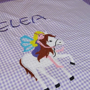 Könnte beinhalten: Eine lila-weiße Gingham-Babydecke mit dem Namen "ELEA" bestickt. Ein weißes Pferd mit einer Fee, die auf seinem Rücken reitet, ist ebenfalls auf die Decke gestickt.