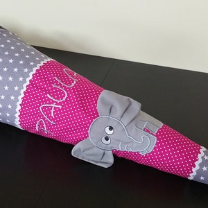 Könnte beinhalten: Eine graue und rosa Schultüte mit weißen Sternen und einem niedlichen Elefanten-Applikation. Auf der Tüte ist der Name "Paula" gestickt und mit einem rosa Band gebunden. Ein lustiges Accessoire für die Einschulung.