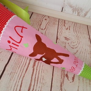 Könnte beinhalten: Ein rosa-grüner Stoffkegel mit einem Hirsch-Motiv, dem Namen "Mila" und dem Datum "09.2017".