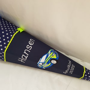 May include: A blue and white school cone with a yellow trim and white stars. The cone has an embroidered police car and the name "Hansen" and the text "Schulkid 2022".