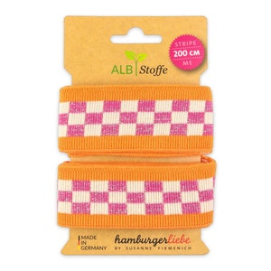 Könnte beinhalten: Orange-weiß kariertes Gummiband mit pinkfarbenem Glitzer, 200 cm lang. Das Gummiband ist auf einem Karton verpackt mit dem Text "ALB Stoffe" und "hamburgerliebe by Susanne Firmenich".