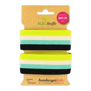 Puede incluir: Dos rollos de cinta elástica a rayas de color verde neón, blanco, turquesa y negro, cada uno de 200 cm. La cinta está empaquetada en un soporte de cartón con el texto "ALB Stoffe" y "hamburgerliebe by SUSANNE FIRMENICH".