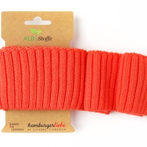 Könnte beinhalten: Orangefarbener gerippter Strickbund, hergestellt in Deutschland von hamburgerliebe, einer Marke von Susanne Firmenich. Der Bund befindet sich auf einem braunen Kartonanhänger mit einem grünen Pflanzenlogo und dem Text "ALB Stoffe".