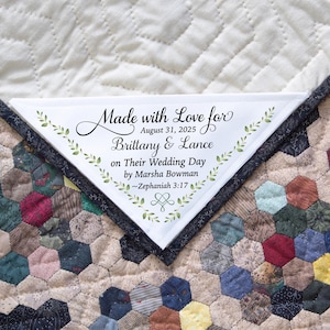 May include: A handmade quilt with a white triangular label. The label reads "Made with Love for Brittany & Lance on Their Wedding Day by Marsha Bowman." The quilt features a patchwork design with various fabric patterns and colors.
