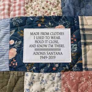 May include: A quilted patchwork blanket featuring a white rectangular fabric label with black text that reads: MADE FROM CLOTHES I USED TO WEAR. HOLD IT CLOSE, AND KNOW I'M THERE. ADONIS SANTANA 1949-2019. The quilt has various floral and plaid patterns.