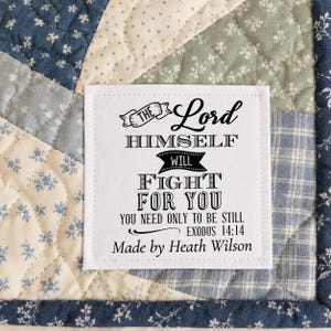 May include: A white fabric label with black text stitched onto a blue, cream, and sage green patchwork quilt. The label reads: THE LORD HIMSELF WILL FIGHT FOR YOU YOU NEED ONLY TO BE STILL EXODUS 14:14 Made by Heath Wilson in black ink.