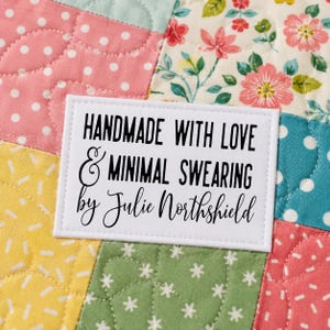 May include: A rectangular white fabric label with black text reading HANDMADE WITH LOVE & MINIMAL SWEARING by Julie Northshield, stitched onto a multicolored quilted fabric featuring pink polka dots, floral patterns, and yellow dashes.