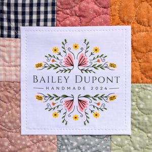 May include: A white square label with the text "BAILEY DUPONT HANDMADE 2024" in gray. The label is decorated with pink and yellow floral designs. The label is stitched onto a patchwork quilt.