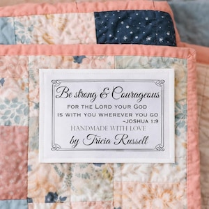 May include: A handmade quilt with a label that reads "Be strong & Courageous." The quilt features a patchwork design with floral patterns in shades of pink, blue, and white, framed by a pink border.