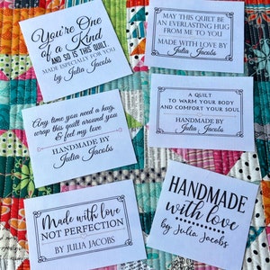 Puede incluir: Varias etiquetas de tela blanca con texto negro se muestran en un edred&oacute;n colorido. Las etiquetas presentan frases como "Hecho a mano con amor" y "Hecho especialmente para ti por Julia Jacobs."