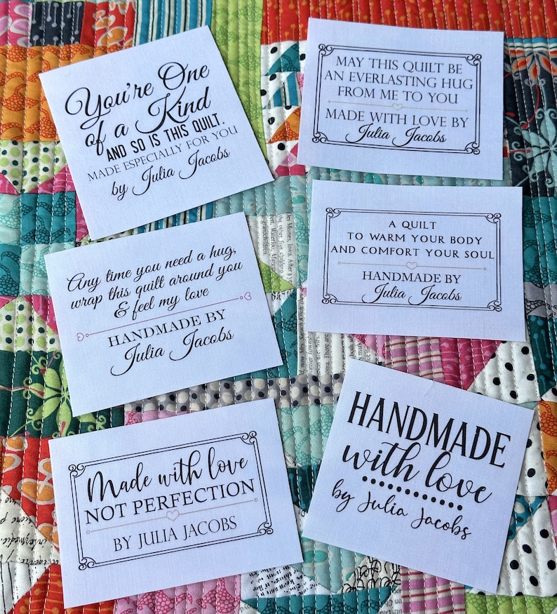 Puede incluir: Varias etiquetas de tela blanca con texto negro se muestran en un edred&oacute;n colorido. Las etiquetas presentan frases como "Hecho a mano con amor" y "Hecho especialmente para ti por Julia Jacobs."