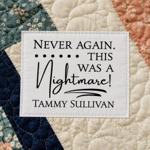 May include: A white rectangular fabric label stitched onto a quilted textile with a floral pattern. The label features black printed text that reads: NEVER AGAIN. THIS WAS A Nightmare! TAMMY SULLIVAN. The label is centered on the fabric background.