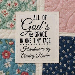 May include: A square fabric label with the text "ALL OF God's GRACE IN ONE TINY FACE Handmade by Ansley Rocha" in black script. The label is on a patchwork quilt with floral and dotted patterns in shades of blue, pink, and green.