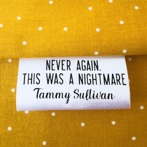 May include: A white label with black text that reads "NEVER AGAIN. THIS WAS A NIGHTMARE Tammy Sullivan" is placed on a yellow fabric with white polka dots. The label is centered and the text is easy to read.