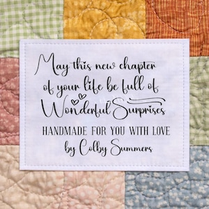 May include: A white fabric label with black script on a patchwork quilt. The text reads "May this new chapter of your life be full of Wonderful Surprises. Handmade for you with love by Colby Summers."