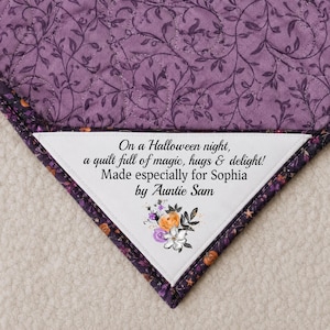 May include: A purple quilt with a floral pattern and a white label. The label reads "On a Halloween night, a quilt full of magic, hugs & delight! Made especially for Sophia by Auntie Sam."