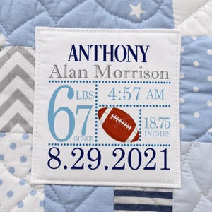 May include: A white fabric square patch featuring the name Anthony Alan Morrison, birth details including 6 lbs 7 ounces, 4:57 AM, 18.75 inches, and the date 8.29.2021, with a brown football graphic, stitched onto a blue, white, and gray patchwork quilt.