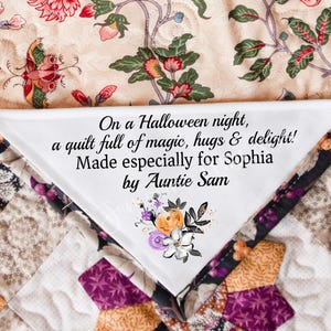 May include: A handmade quilt with a white label featuring text: "On a Halloween night, a quilt full of magic, hugs & delight! Made especially for Sophia by Auntie Sam." The label has a floral design with orange, purple, and white flowers. The quilt has a patchwork design.