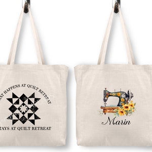 May include: Two white canvas tote bags with different designs. The first bag has a black and white quilt block design with the text "What happens at quilt retreat stays at quilt retreat". The second bag has a floral decorated sewing machine with the text "Marin".