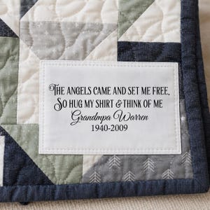 May include: A quilted fabric patch in white with black text that reads: The angels came and set me free, so hug my shirt & think of me, Grandpa Warren, 1940-2009. The patch is stitched onto a patchwork quilt featuring navy blue, sage green, and white fabric.