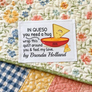 May include: A white square patch with the text "IN QUESO you need a hug wrap this quilt around you & feel my love. by Brenda Holland" and a graphic of a tortilla chip dipping into a bowl of cheese. The patch is on a floral quilt.
