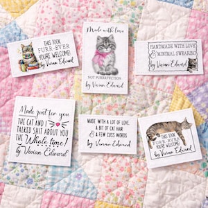 May include: A collection of white fabric labels with cat-themed illustrations and text. The labels feature phrases like "Made with love," "Not purrrfection," and "This took furr-ever." The labels are designed by Vivian Edward.
