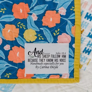 May include: Close-up of a handmade quilt with a floral pattern in orange, yellow, and pink on a blue background. The quilt has a yellow border and a white label with text from John 10:4 and the maker's name.