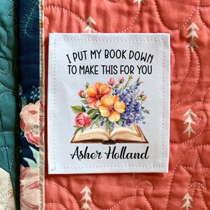 May include: White fabric square with a stitched border. The square has a floral design with a book and the text "I put my book down to make this for you Asher Holland".