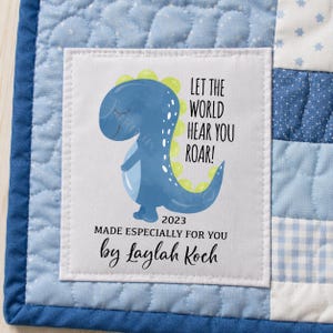 May include: A blue and white baby quilt with a central square featuring a blue dinosaur illustration and the text "LET THE WORLD HEAR YOU ROAR!". The text "MADE ESPECIALLY FOR YOU by Laylah Koch" is also included.