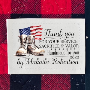May include: A white fabric label with a thank you message for service, sacrifice, and valor. The label features a watercolor illustration of a pair of brown boots and an American flag. The label also includes the text "Handmade for you" and "by Makaila Robertson".