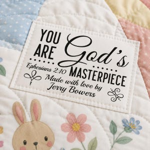May include: A rectangular white fabric label stitched onto a quilted blanket. The label features black text reading "YOU ARE God's MASTERPIECE Ephesians 2:10 Made with love by Jerry Bowers" with decorative flourishes on a background of pastel polka dots.