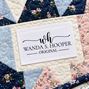 May include: A close-up of a quilt with a white label that reads "wh WANDA S. HOOPER ORIGINAL" in black script. The quilt features a patchwork design with blue, pink, and white floral patterns and geometric shapes.