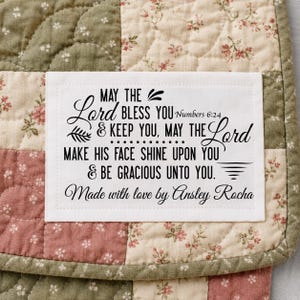 May include: Close-up of a patchwork quilt with a white label. The label features black script with the text "MAY THE Lord BLESS YOU & KEEP YOU, MAY THE Lord MAKE HIS FACE SHINE UPON YOU & BE GRACIOUS UNTO YOU. Made with love by Ansley Rocha."