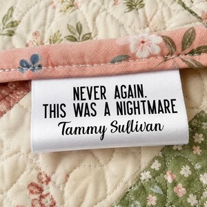 May include: A white rectangular fabric label sewn onto a quilted floral fabric with a pink border. The label features black text that reads: NEVER AGAIN. THIS WAS A NIGHTMARE, followed by the name Tammy Sullivan in a cursive script font.