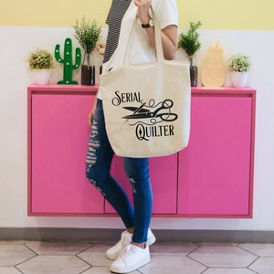 May include: A white canvas tote bag with black text and a graphic of scissors and a needle that reads "Serial Quilter".
