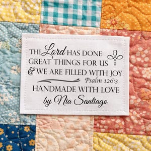 May include: A white rectangular label with black text, stitched onto a patchwork quilt. The text reads "THE Lord HAS DONE GREAT THINGS FOR US & WE ARE FILLED WITH JOY Psalm 126:3 HANDMADE WITH LOVE by Nia Santiago."
