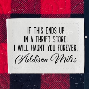 May include: A white rectangular label with the text "IF THIS ENDS UP IN A THRIFT STORE, I WILL HAUNT YOU FOREVER. Addison Miles" in black. The label is set against a red and black plaid background.