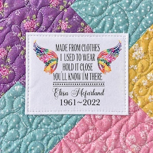 May include: A white fabric label with the text "MADE FROM CLOTHES I USED TO WEAR HOLD IT CLOSE YOU'LL KNOW I'M THERE" and the name "Elisa Mcfarland 1961~2022". The label has colorful butterfly wing designs and is set on a quilted background.
