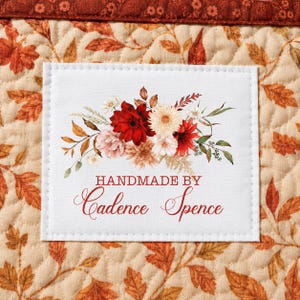 May include: A white rectangular fabric label featuring a floral illustration of red, pink, and cream flowers with green leaves. The text "HANDMADE BY Cadence Spence" is printed in red. The label is stitched onto a quilted fabric with autumn leaves.