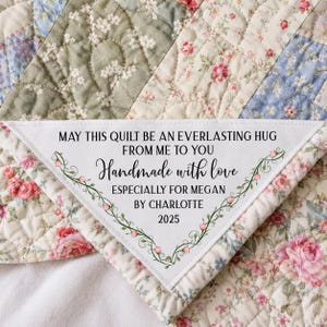 May include: A quilted fabric piece featuring a floral pattern in shades of pink, blue, and green. A white triangular patch is sewn onto the quilt with the text: MAY THIS QUILT BE AN EVERLASTING HUG FROM ME TO YOU Handmade with love ESPECIALLY FOR MEGAN BY CHARLOTTE 2025, framed by green vines and small pink flowers.