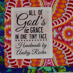 May include: White fabric label with black text that reads "All of God's Grace in one tiny face... Handmade by Ansley Rocha". The label is on a colorful fabric background.