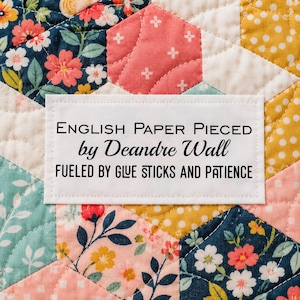 May include: A white rectangular fabric label stitched onto a multicolored floral quilt. The label features black text reading: ENGLISH PAPER PIECED by Deandre Wall FUELED BY GLUE STICKS AND PATIENCE. The quilt has a hexagonal patchwork pattern.