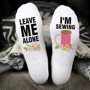 May include: A pair of white socks with a fun design. One sock says "Leave Me Alone" with a floral design. The other sock says "I'm Sewing" with a pink spool of thread and needle design.